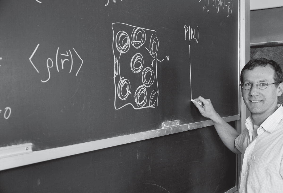 Professor Phillip Geissler looking at camera, in black and white at chalkboard drawing line with chalk